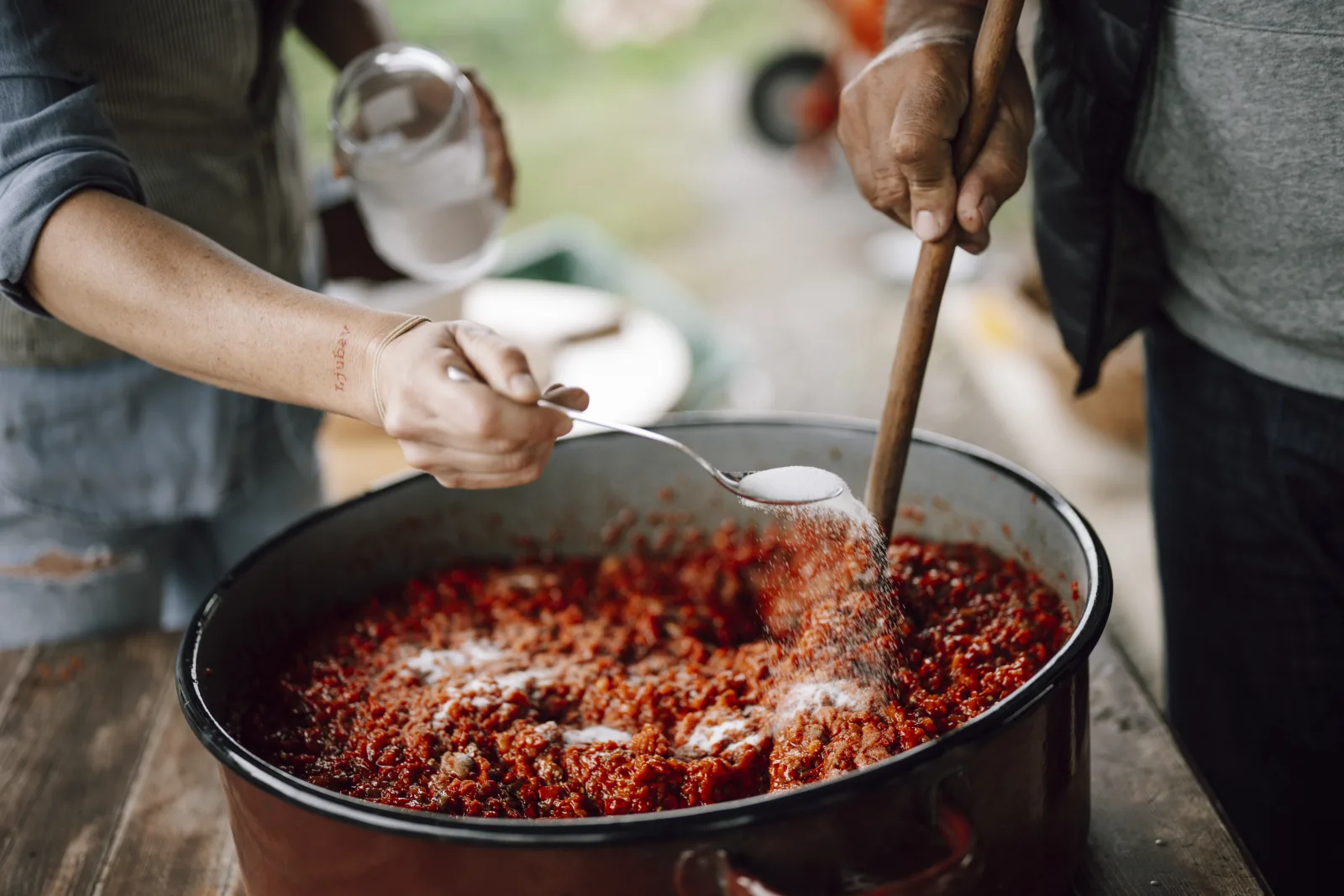 In a rustic outdoor kitchen, two people prepare a flavorful dish. One stirs a large pot filled with colorful ingredients, adding seasonings with care amidst a natural setting.