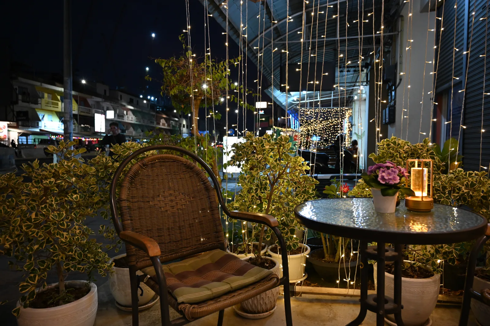 This photo shows a cozy nighttime balcony or terrace setting in Pattaya City, Thailand. There's a comfortable wicker chair with a cushion in the foreground, positioned next to a small round table that has a candle lantern and what appears to be a potted flower on it. The space is beautifully decorated with string lights hanging vertically, creating a curtain-like effect that adds a warm, magical ambiance. Several potted plants and small shrubs surround the seating area, adding greenery to the space. In the background, you can see city lights and buildings, suggesting this is a rooftop terrace or balcony with an urban view. The overall atmosphere is intimate and relaxing - perfect for evening relaxation or dining while enjoying the city lights.