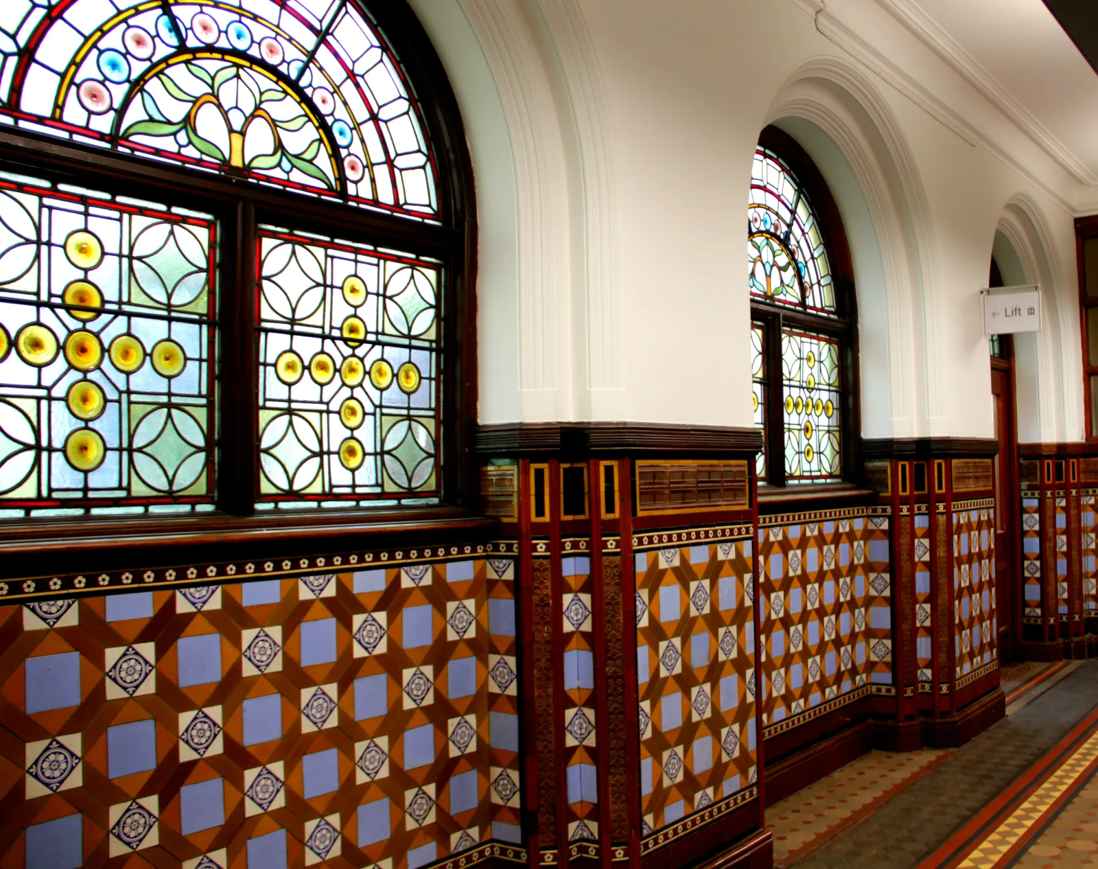 Interior of the former Municipal Buildings (now the Central Library), Leeds, West Yorkshire, England, built 1879-1884 to the design of George Corson. 
The general interior carving was executed by a team led by John Wormald Appleyard. The alabaster arch in the main entrance was made by Farmer &amp; Brindley of London, and the reading room (now cafe) medallions were carved by Benjamin Creswick. George Wilson made the leaded clerestory windows of the original reference library, and Powell Bros made the rest of the stained glass and leaded lights. Doulton supplied the terracotta tiles in what is now (2019) the art library. Ceramic wall and floor tiles were supplied by E. Smith of Coalville, Leicester, and J. Taylor of Leeds. Curtis &amp; Son made the bookcases and mirror screens in the gallery of the original reference library (as of 2019 the family history department). Jones of Manchester made the wrought-iron gates and railings outside, but Smith &amp; Sons of Birmingham made the entrance gates, interior grilles and original door furniture (only a few of these last items remain). The interior was originally decoratively painted, in gold and colours, by J.T. Pollard of Leeds.

Showing corridor on an upper floor.