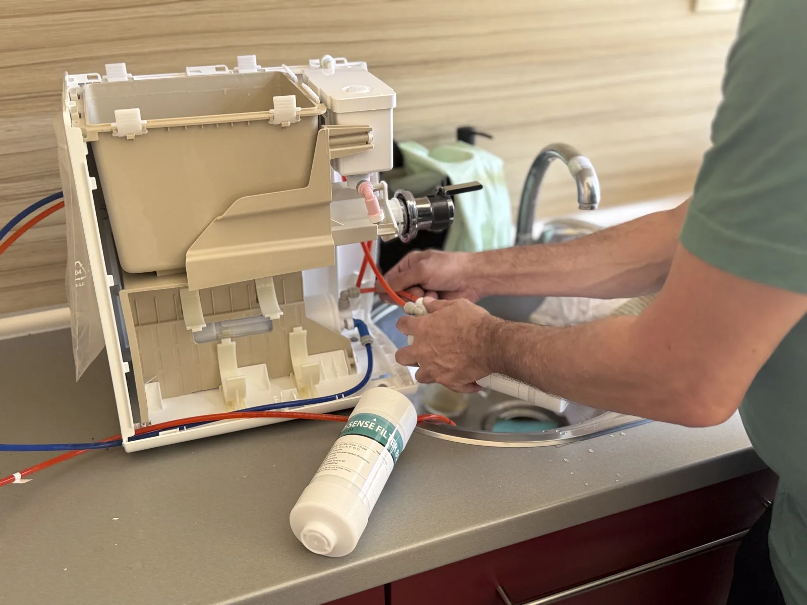 A person is working to fix a kitchen appliance at a home kitchen sink. The counter has tools and a cleaning bottle. Water is seen flowing near the appliance.