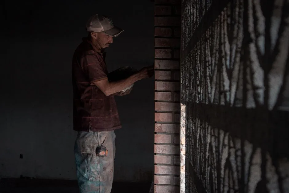 A construction worker laying bricks inside a dimly lit room, illustrating dedication.