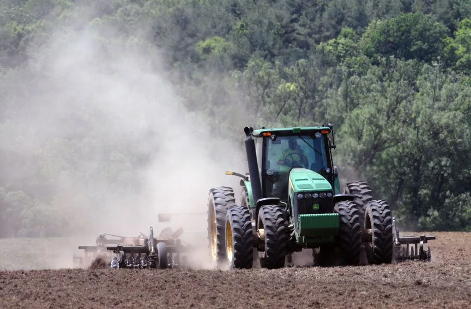 A green tractor plowing a field, creating clouds of dust in an agricultural setting.