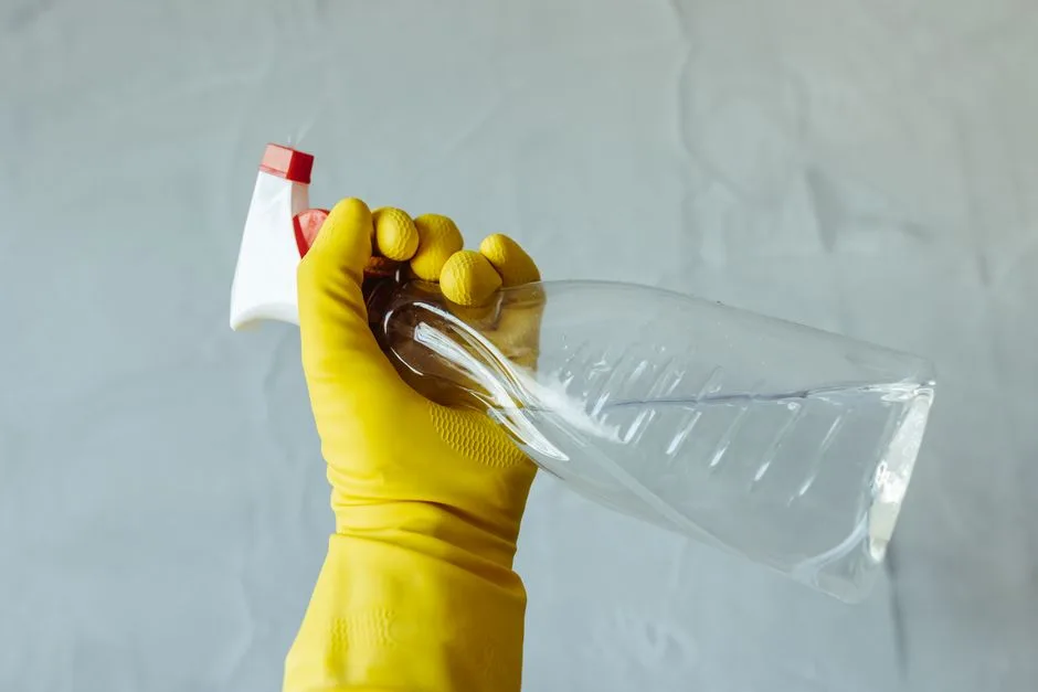 Close-up of a hand wearing yellow gloves holding a transparent spray bottle, isolated on a plain background.