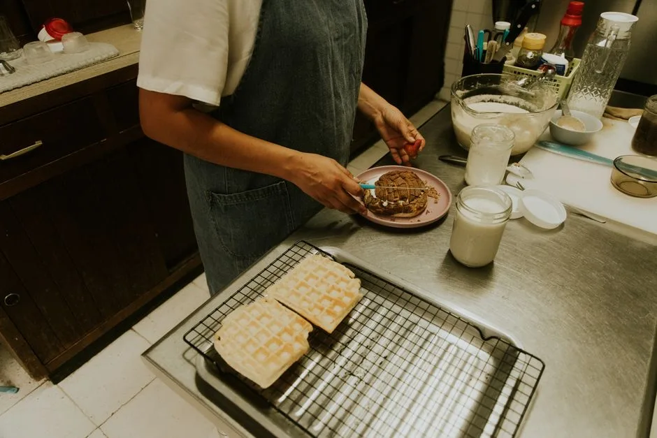 A woman in a denim apron prepares waffles in a home kitchen setting, adding a personal touch.