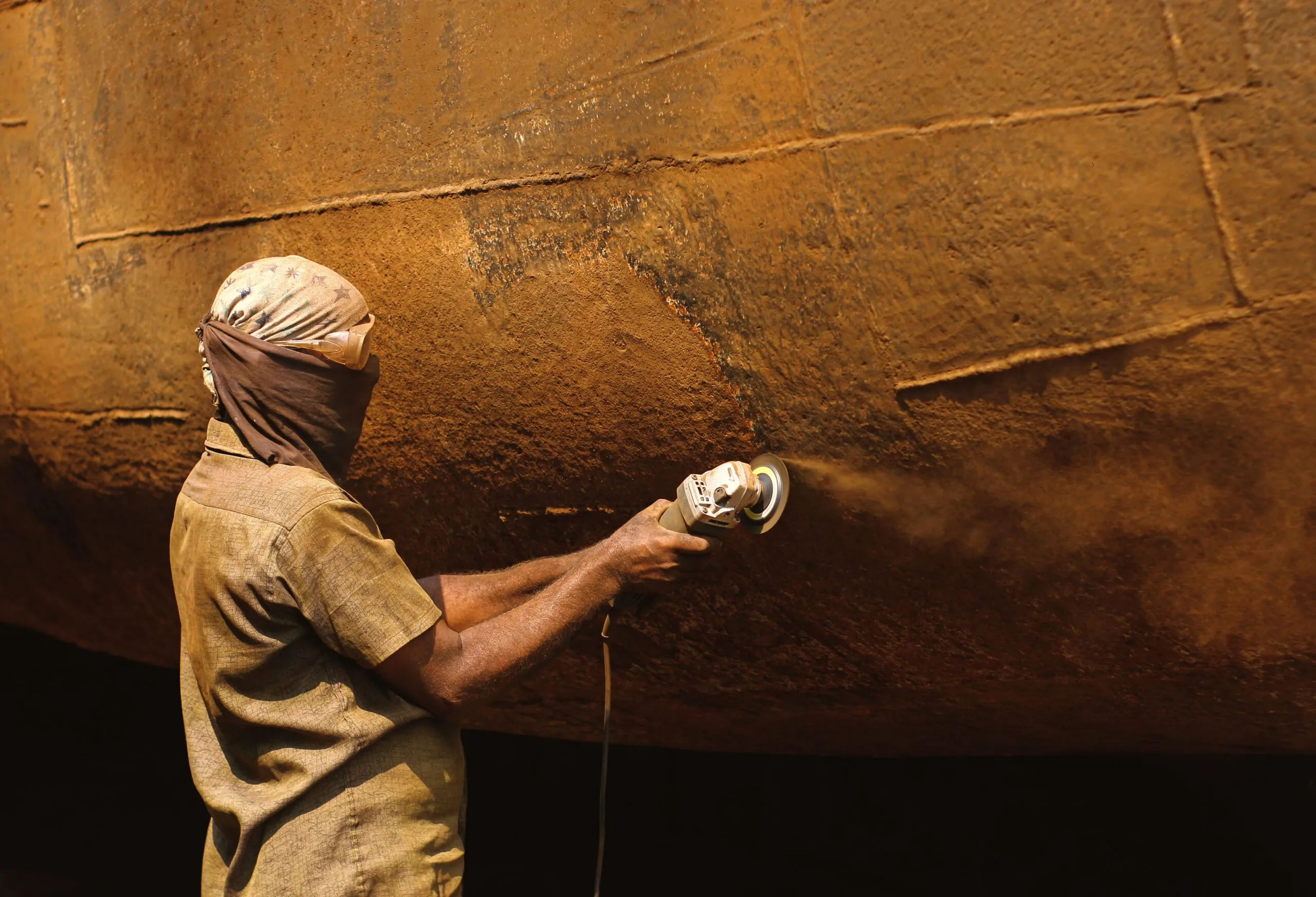 Industrial worker grinding rust off metal surface in outdoor setting.