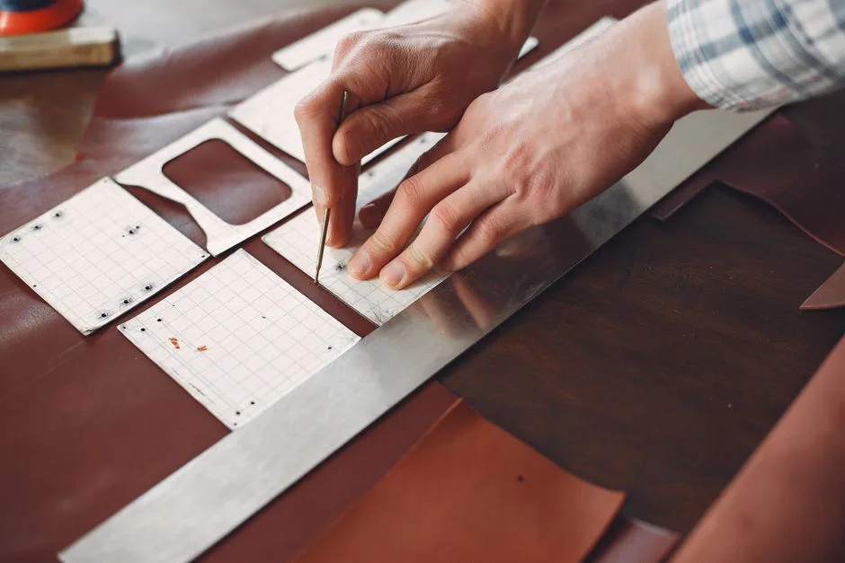 From above of faceless craftsman using ruler and pattern and cutting leather strip for belts while working in modern workshop