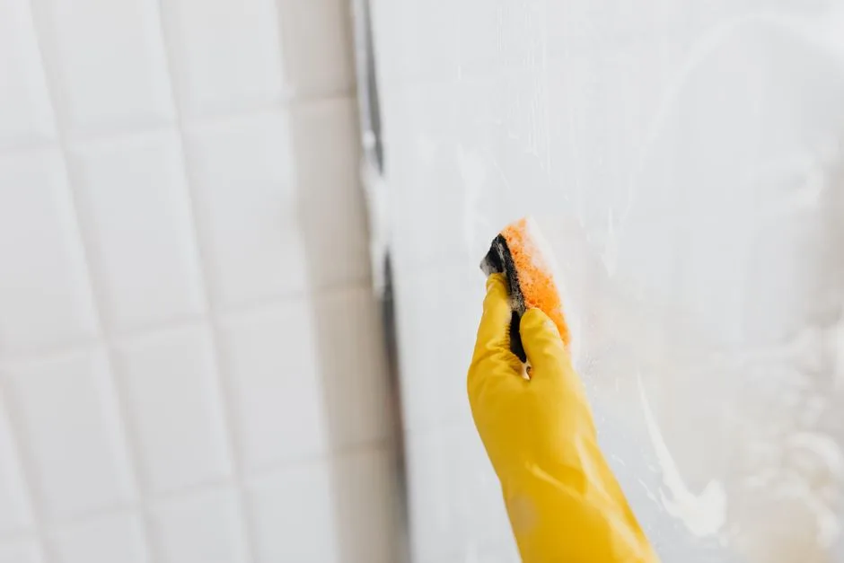 From above of crop anonymous person in yellow rubber protective glove washing shower cabin glass with sponge and detergent