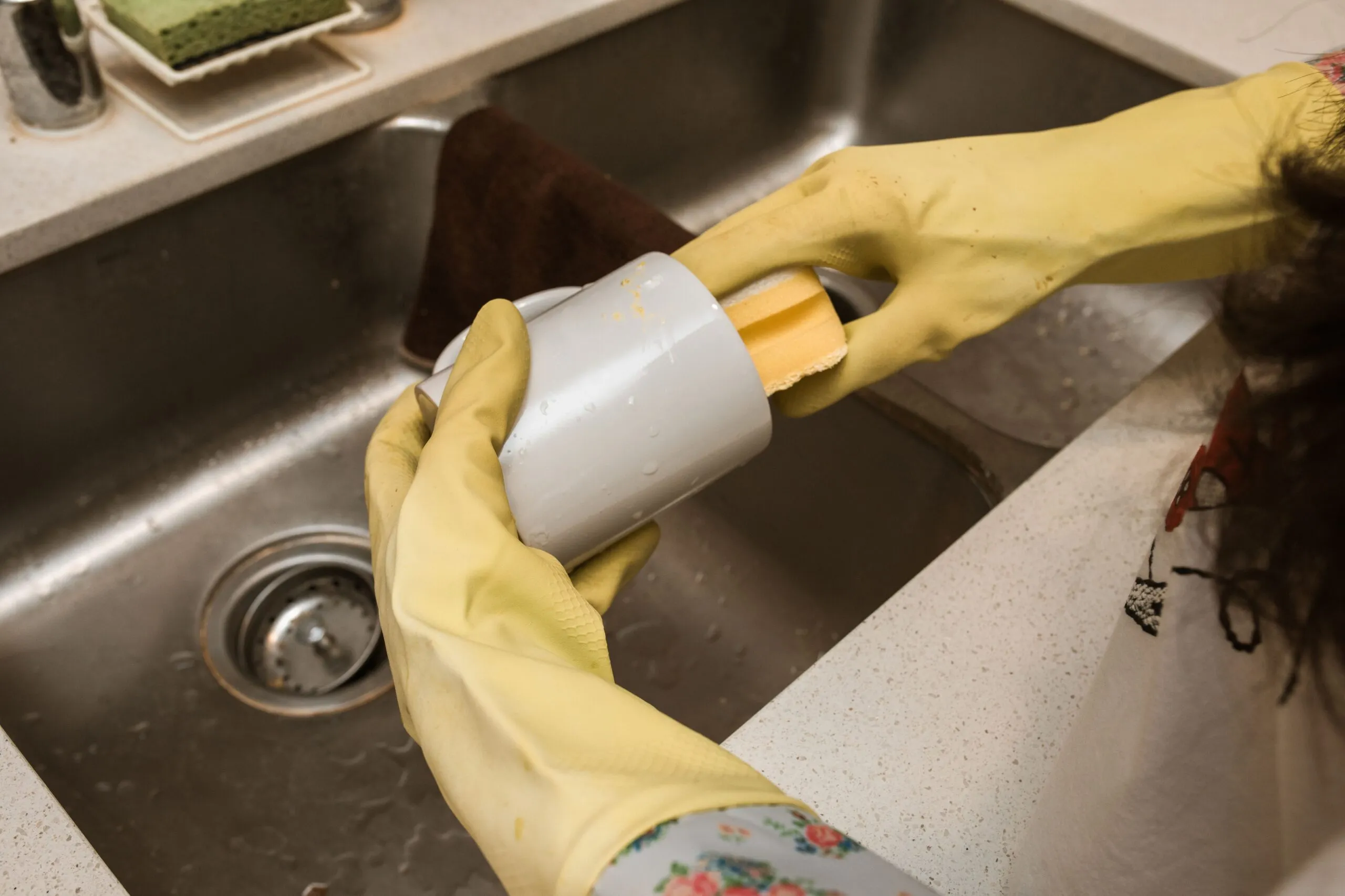 Close-up of a person washing a mug with yellow rubber gloves and sponge in a kitchen sink.