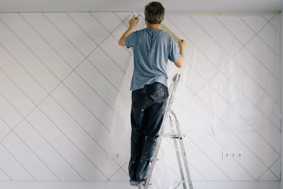 Full body back view of faceless male in shirt and dirty pants repainting wall while standing on ladder in bright empty room in daytime