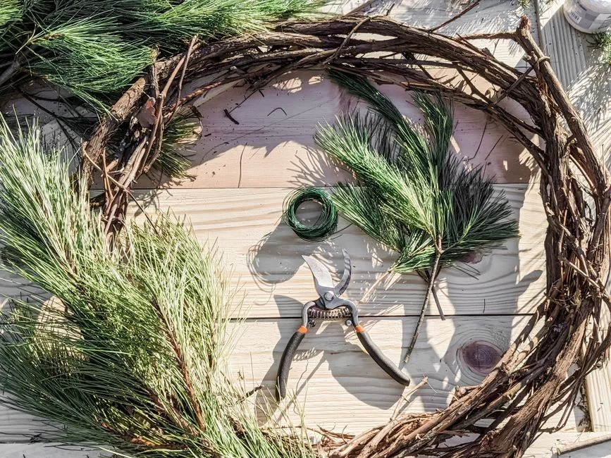 Top view of a DIY pine wreath with tools and branches on a wooden table.