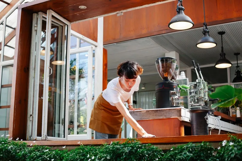 Young waitress in casual clothes and apron standing in modern cafeteria and cleaning wooden bench in daytime