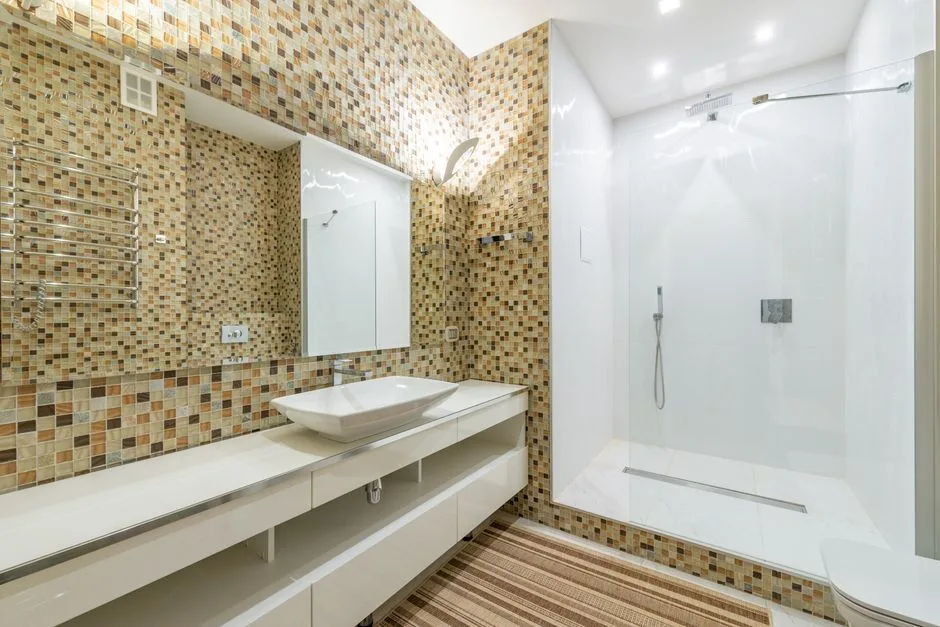 Wide angle of interior of contemporary bathroom with shower cabin and large mirror above white clean sink at mosaic tiled wall