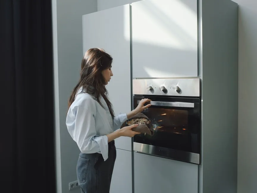 Young woman baking at home, placing a dish in the oven. Bright natural lighting in a modern kitchen setting.