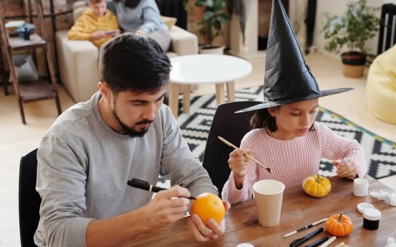 A father and daughter painting pumpkins together indoors, celebrating Halloween creativity.