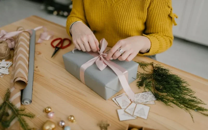 Woman in yellow sweater wrapping a gift with pink ribbon on wooden table decorated for Christmas.