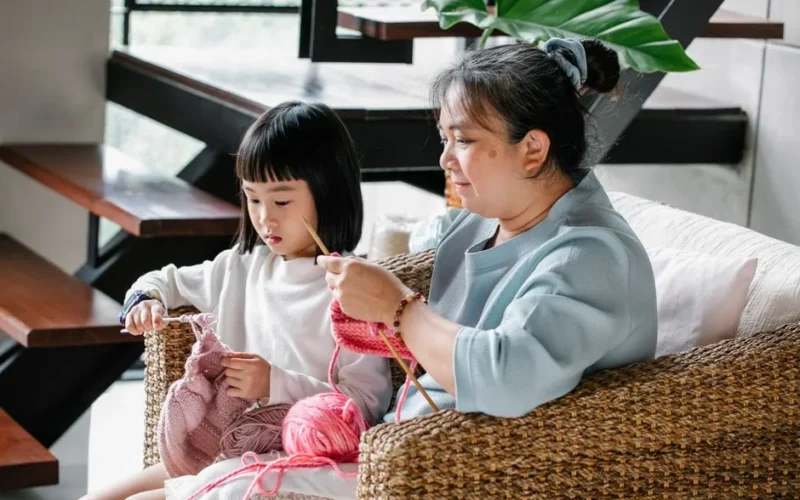 Little Asian girl with mature grandmother sitting in armchair and knitting with needles at home