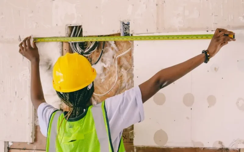 Female construction worker with hardhat and vest measuring an interior wall.