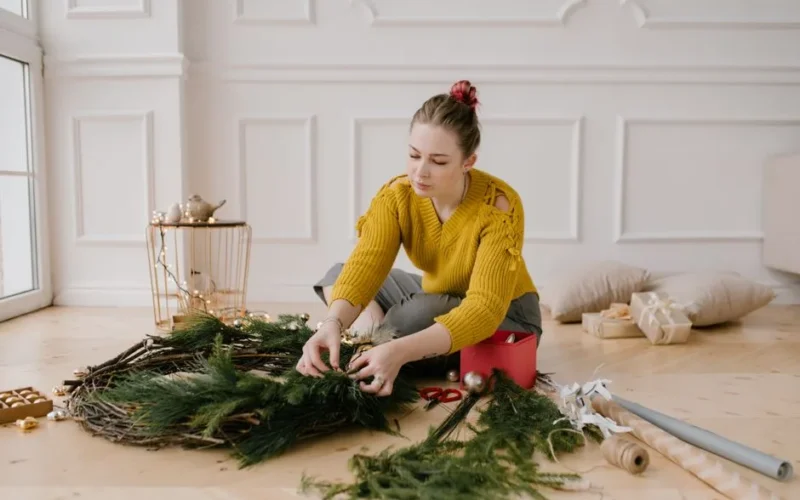 Woman decorating a Christmas wreath inside a cozy room, focused on holiday preparations.