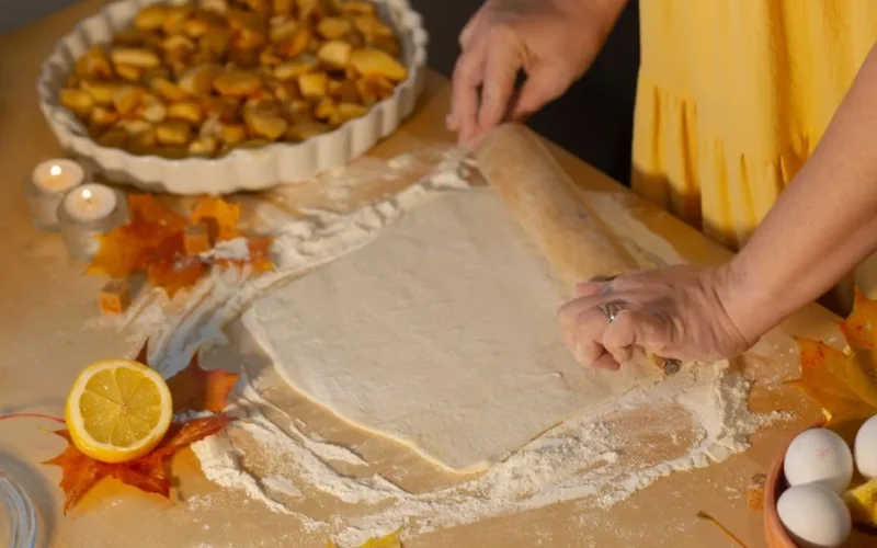 Woman rolling dough for apple pie surrounded by autumn decor and ingredients.