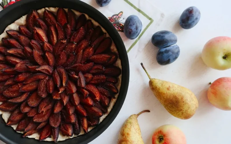 A delicious plum tart surrounded by fresh autumn fruits on a white table.