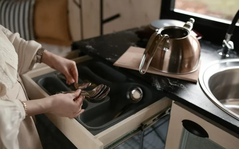 A woman organizing spoons in a kitchen drawer beside a stainless steel kettle.