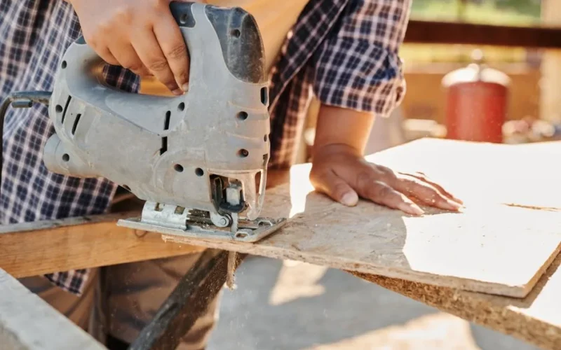 Carpenter using a jigsaw to cut plywood at a construction site, showcasing woodworking skill.