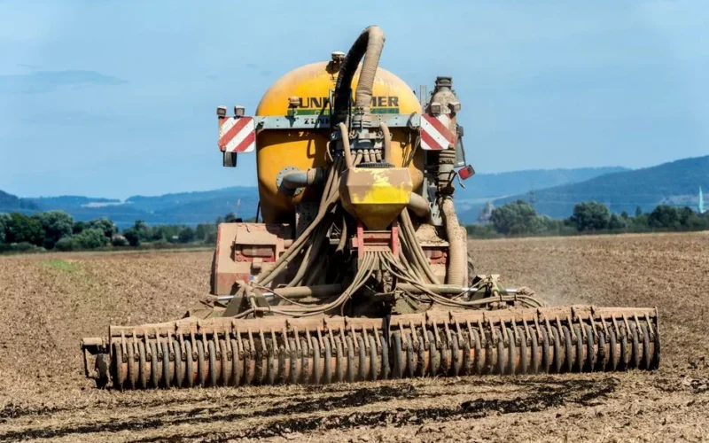 A rear view of farm machinery fertilizing a field in the countryside under a clear blue sky.