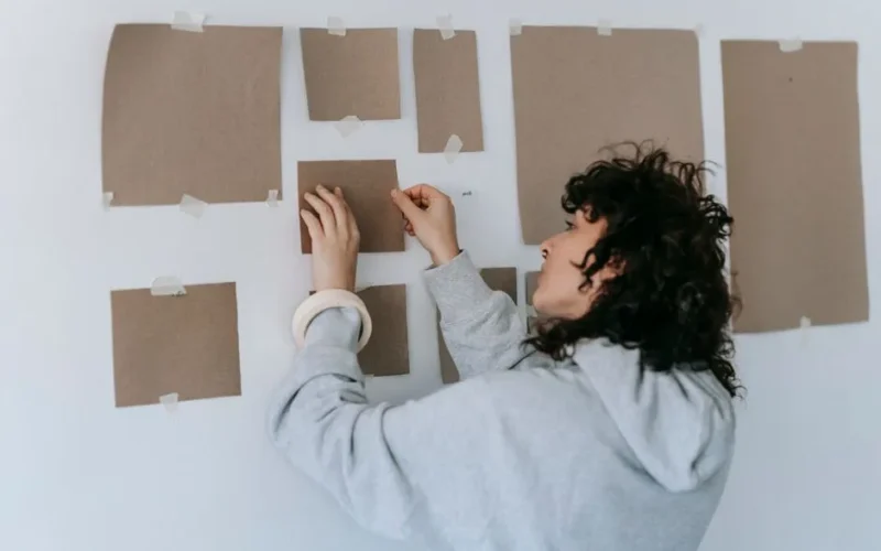 A woman in casual attire arranges cardboard pieces on a wall during an indoor art project.