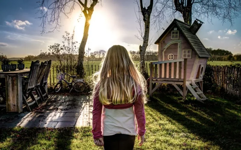 A child enjoying a sunny day in a backyard with a wooden playhouse and trees.