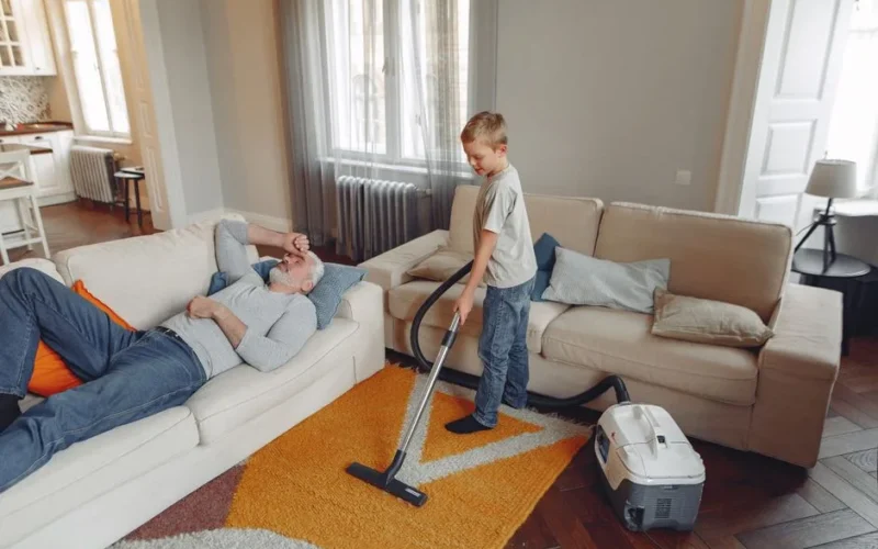 A young boy vacuums the living room carpet while his grandfather relaxes on the sofa.