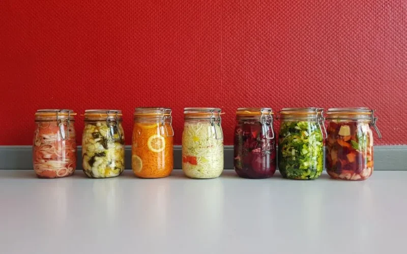 A row of vibrant, multicolored mason jars filled with assorted fermented vegetables against a red wall.