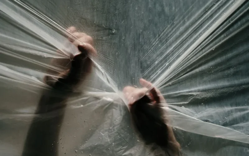 Hands gripping and stretching a transparent plastic film with water droplets and light effects.