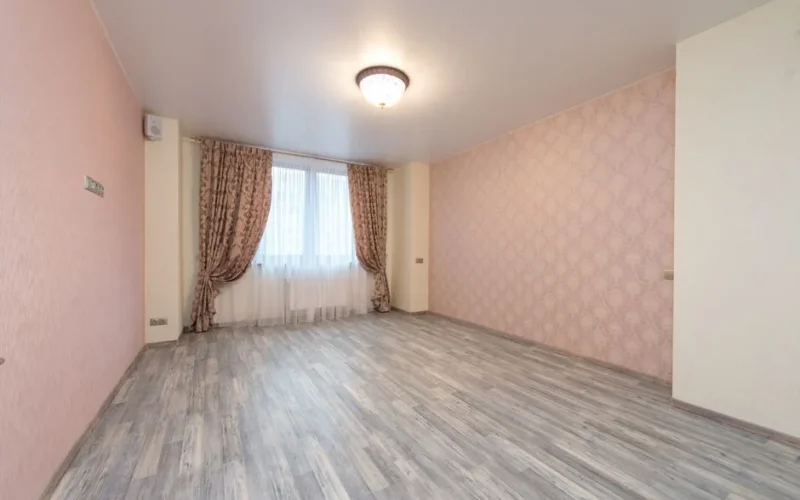 Empty spacious room with laminate and window with colorful curtains between walls with pink ornamental wallpapers and glowing white chandelier