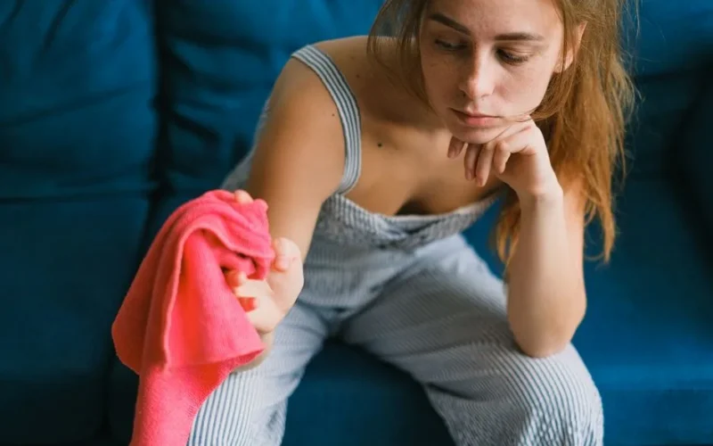 Crop exhausted young female with long hair in casual clothes leaning on hand while sitting on couch with dusting cloth during household routine