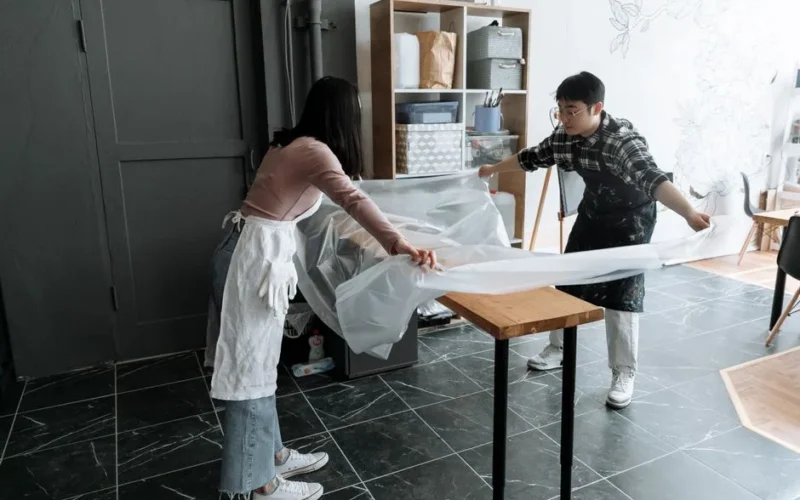 Two adults setting up a workspace with aprons and plastic sheet indoors.