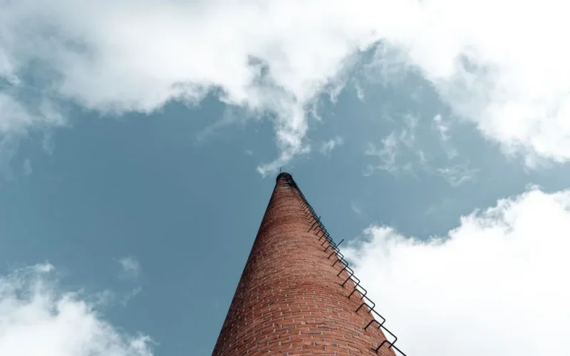 A towering brick chimney with a ladder reaching into a blue sky scattered with white clouds, captured from a low angle.