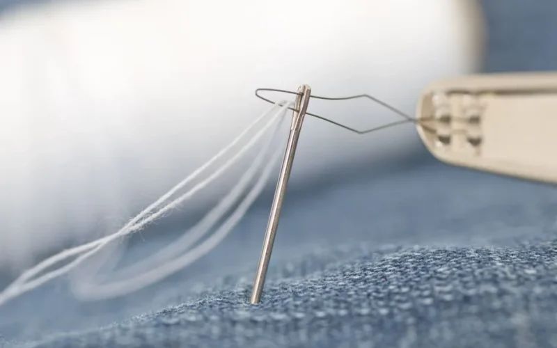 Detailed macro shot of a sewing needle threading white thread through denim fabric.