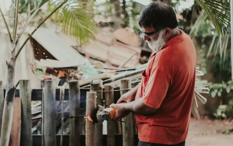 An adult man wearing a red shirt uses a grinder on a wooden fence with sparks flying, showcasing outdoor work.