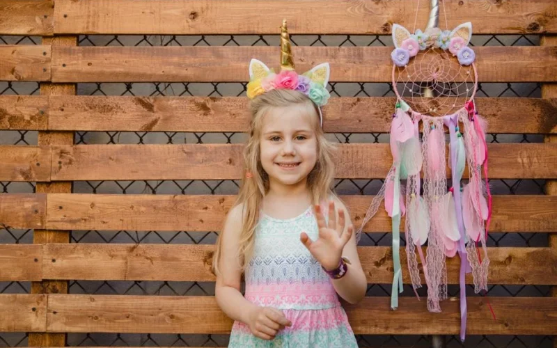 A cheerful girl wearing a unicorn headband stands next to a colorful dreamcatcher against a wooden background.