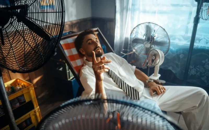 A man in a white suit lounges in a warm room, cooled by multiple fans.