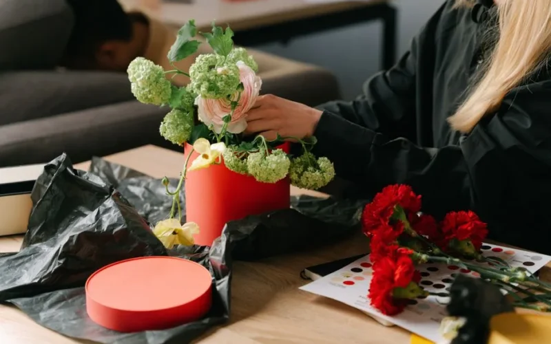 A woman arranges flowers in a red hat box on a wooden table in a cozy indoor setting.