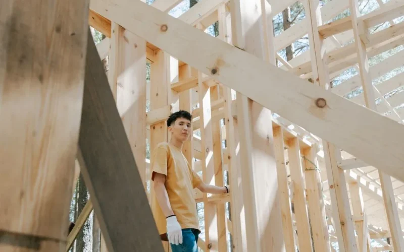 Man in brown shirt and gloves building a wooden structure in sunlight.