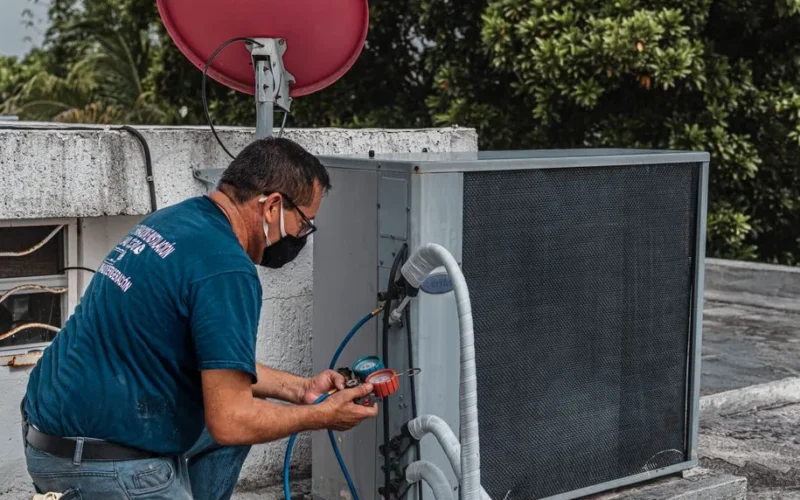 Technician repairing an air conditioner unit outdoors, wearing a facemask and using a manifold gauge.