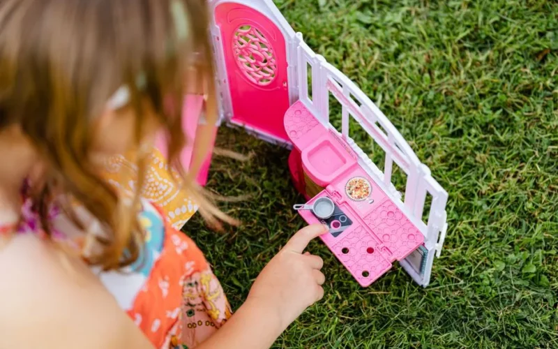 A young girl plays with a pink toy kitchen set on green grass, dressed in a floral dress.