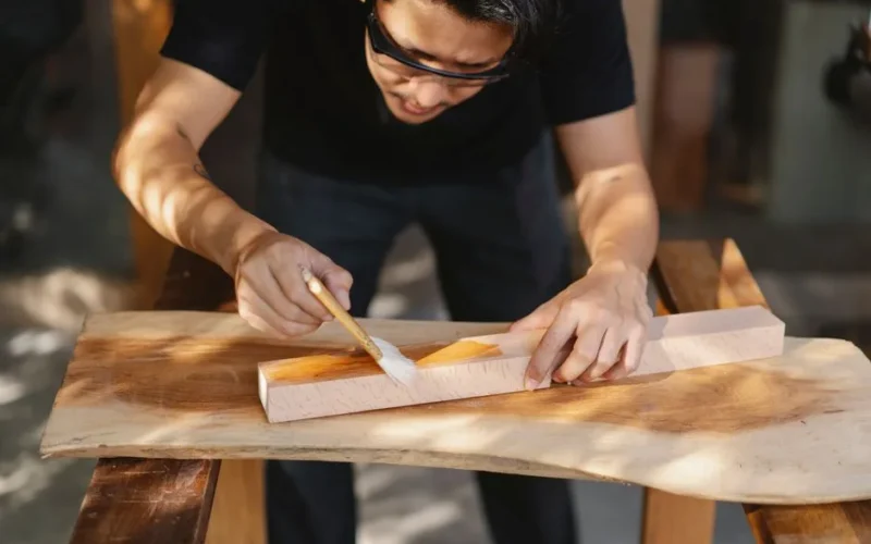 Young ethnic carpenter applying protective varnish on wooden detail while working in professional workshop