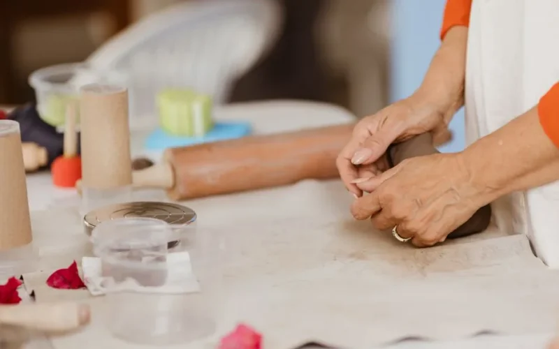 Hands working clay on a table, surrounded by craft tools, indicating a warm creative indoor atmosphere.
