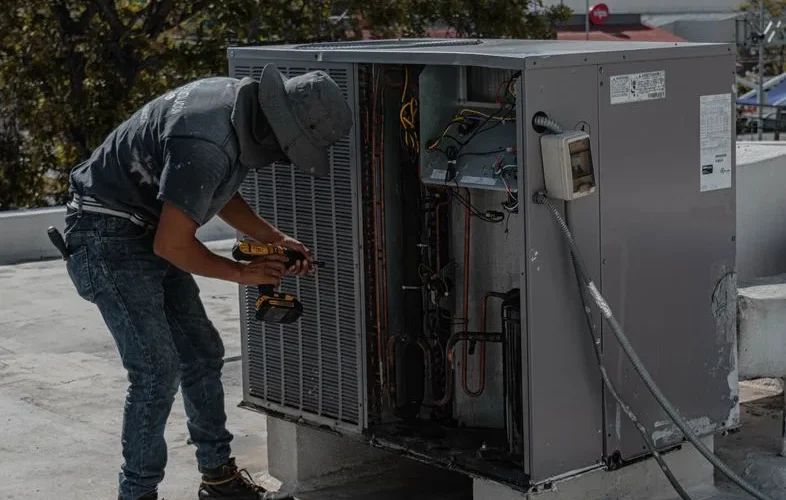 A worker in a bucket hat repairs an outdoor air conditioning unit on a rooftop.