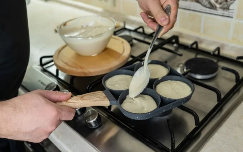 Close-up of a person preparing pancakes on a stovetop using a ladle and batter-filled skillet.