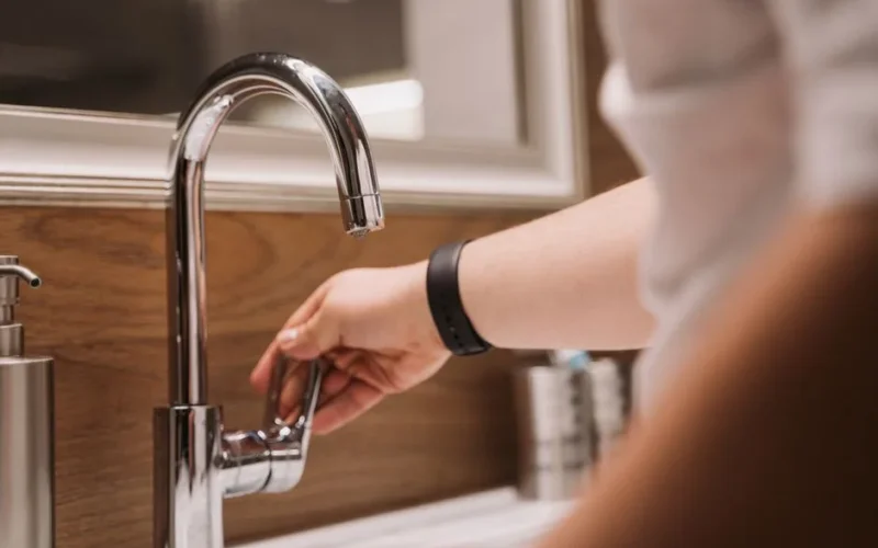 Close-up of a person's hand turning a chrome faucet handle in a modern bathroom setting.