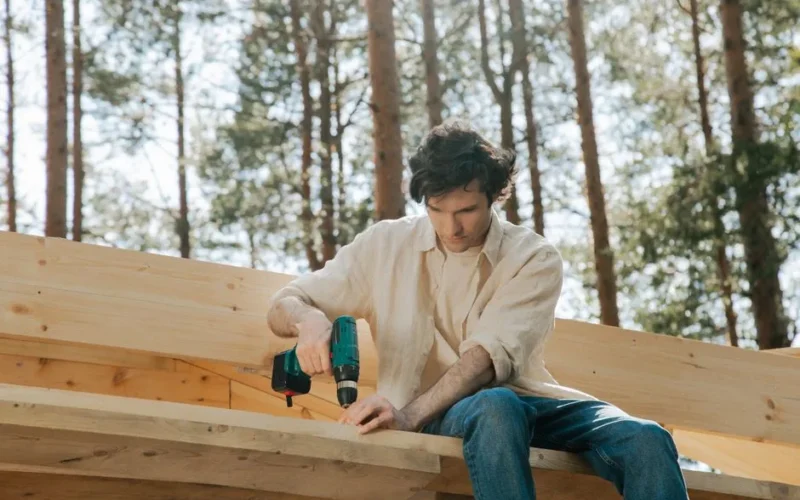Focused carpenter uses power drill on wooden beams in an outdoor setting, surrounded by trees.